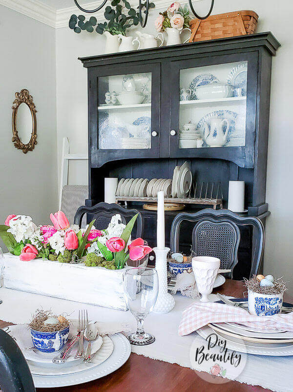 Easter Tablescape ideas, moss and eggs are placed in blue and white teacups at each place setting with white layered plates. White tablerunner with a white washed wooden box filled with white and pink tulips.